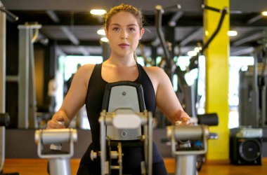 Genç bir kadın, modern bir spor salonundaki bir makinede güç eğitimine odaklanmış. Spor salonunda disiplinli egzersiz yaparak sağlıklı yaşam tarzına ve sağlığına kendini adamış.