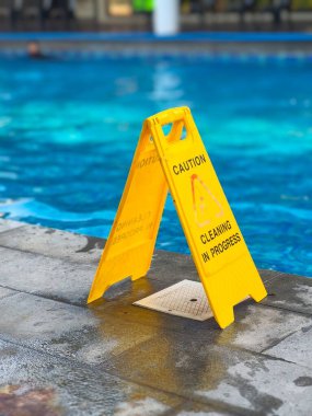 Yüzme havuzunun yakınındaki ıslak havuz kenarında sarı uyarı işareti var. Temizlik yapılmakta ve güvenlik açısından kaygan zemin tehlikesi uyarısı var..