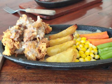 Sıcak bir tavada servis edilen leziz kızarmış tavuk, kremalı sos, patates parçaları, mısır, havuç ve yeşil fasulye..