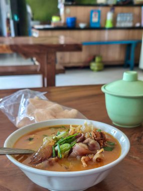 Baharatlı et tendonu çorbası, şifalı otlar ve yeşil soğanlı et suyu. Krakerler ve ahşap masada yeşil bir tabakla birlikte beyaz bir kasede servis edilir..