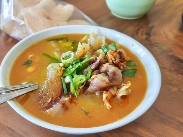 Baharatlı et tendonu çorbası, şifalı otlar ve yeşil soğanlı et suyu. Krakerler ve ahşap masada yeşil bir tabakla birlikte beyaz bir kasede servis edilir..