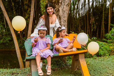 Colombian mother smiling at camera while twin daughters make playful faces with cupcakes during birthday celebration in park