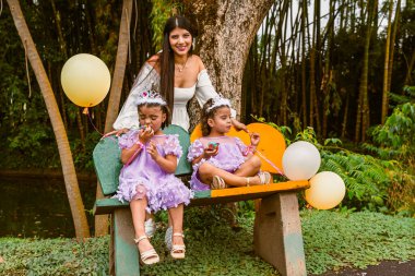 Colombian mother smiling at camera while twin daughters enjoy cupcakes during peaceful birthday moment in lush park