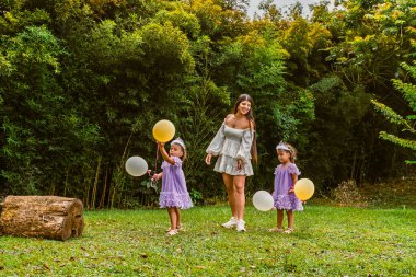 Mother smiles at twin daughters holding balloons during outdoor birthday, as one girl looks upset
