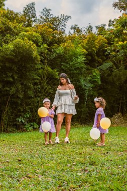 Mother holds hands with twin daughters during outdoor birthday, as one girl shows angry expression