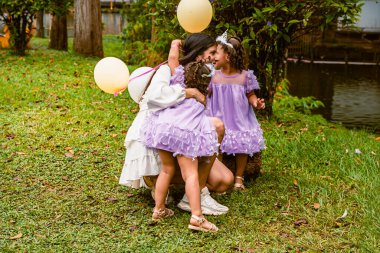 Mother hugs twin daughters in lavender dresses during outdoor birthday celebration as sibling watches with balloon