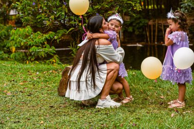 Mother hugs daughter with balloon while twin sister watches during outdoor birthday celebration in nature