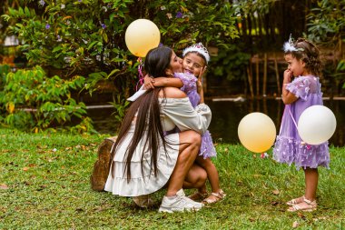 Mother hugs daughter with balloon while twin sister watches during outdoor birthday celebration