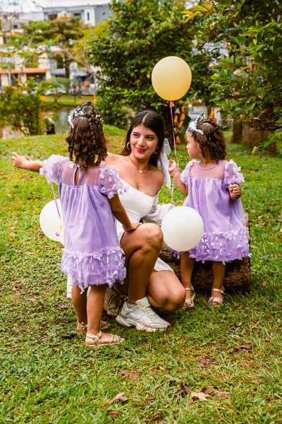 Mother interacts with twin daughters holding balloons during outdoor birthday celebration in nature