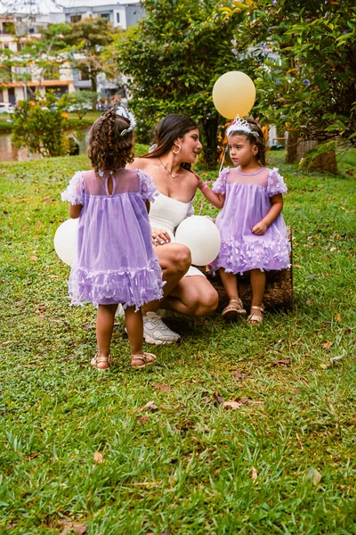Mother interacts with twin daughters in lavender dresses while sibling watches with balloon during outdoor birthday celebration
