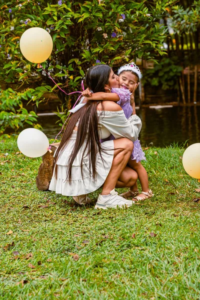 Mother hugs daughter with tiara and balloon near tree during outdoor birthday celebration