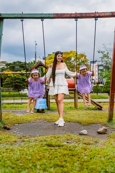 Mother supports twin daughters on swings during outdoor birthday celebration in playground
