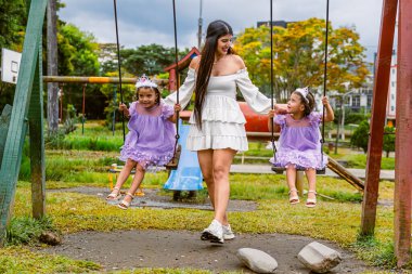 Mother holds hands of twin daughters on swings during outdoor birthday celebration in autumn park
