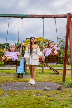 Mother holds hands of twin daughters on swings during outdoor birthday celebration in cloudy playground