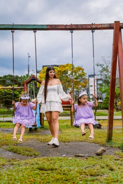 Mother holds hands of twin daughters on swings during outdoor birthday celebration in urban playground