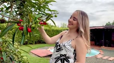 Pregnant Colombian woman smiles while touching red fruits in garden near pool, peaceful rural moment