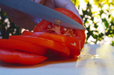 bir kesme tahtası ağaçlar ve günbatımı akşam arka planı üzerinde bir domates el ve bıçak ile dilimleme portre