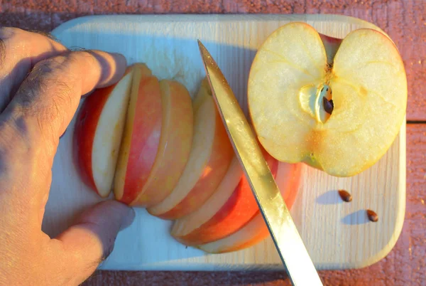 Cutting an apple with his hand with knife on cutting wooden cutting board on a red-brown background at sunset — Stock Photo, Image