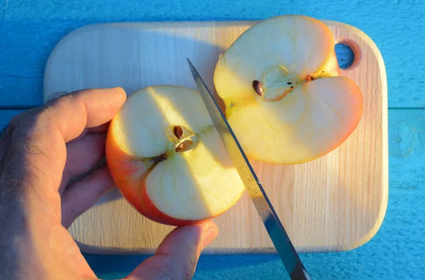 Cutting an apple with his hand with knife on cutting wooden cutting board on a turquoise background at sunset — Stock Photo, Image