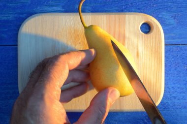 bir el ile bir armut uzun mavi gölgeli mavi ahşap bir masa üzerinde bir ahşap kesme tahtası üzerinde bıçak ile kesme