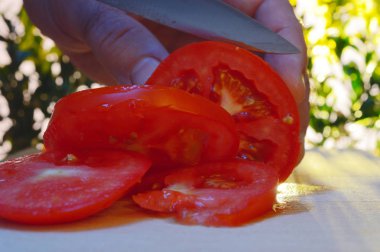 bir kesme tahtası ağaçlar ve günbatımı akşam arka planı üzerinde bir domates bir el ve bıçak ile dilimleme portre