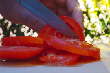 bir kesme tahtası ağaçlar ve günbatımı akşam arka planı üzerinde bir domates bir el ve bıçak ile dilimleme portre