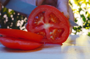 bir kesme tahtası ağaçlar ve günbatımı akşam arka planı üzerinde bir domates bir el ve bıçak ile dilimleme portre