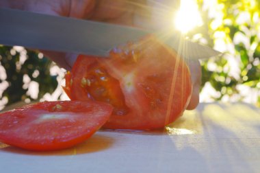 bir kesme tahtası ağaçlar ve günbatımı akşam arka planı üzerinde bir domates bir el ve bıçak ile dilimleme portre