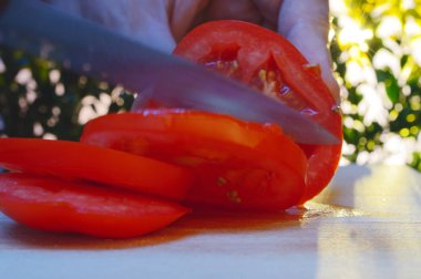 bir kesme tahtası ağaçlar ve günbatımı akşam arka planı üzerinde bir domates bir el ve bıçak ile dilimleme portre