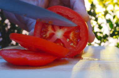 bir kesme tahtası ağaçlar ve günbatımı akşam arka planı üzerinde bir domates bir el ve bıçak ile dilimleme portre