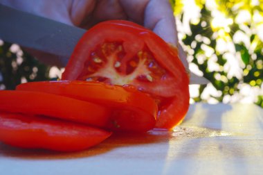 bir kesme tahtası ağaçlar ve günbatımı akşam arka planı üzerinde bir domates bir el ve bıçak ile dilimleme portre