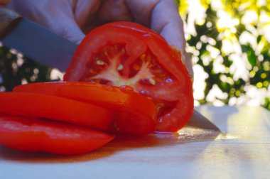 bir kesme tahtası ağaçlar ve günbatımı akşam arka planı üzerinde bir domates bir el ve bıçak ile dilimleme portre