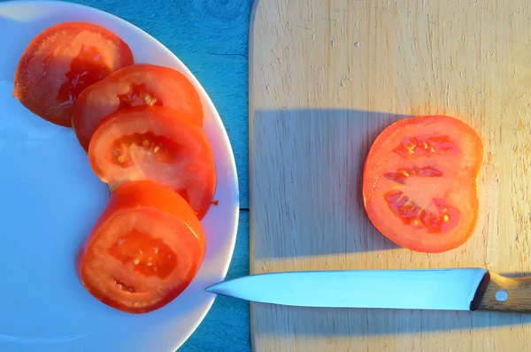 sliced tomato slices on a plate and a wooden cutting board on a blue table lit by the sun