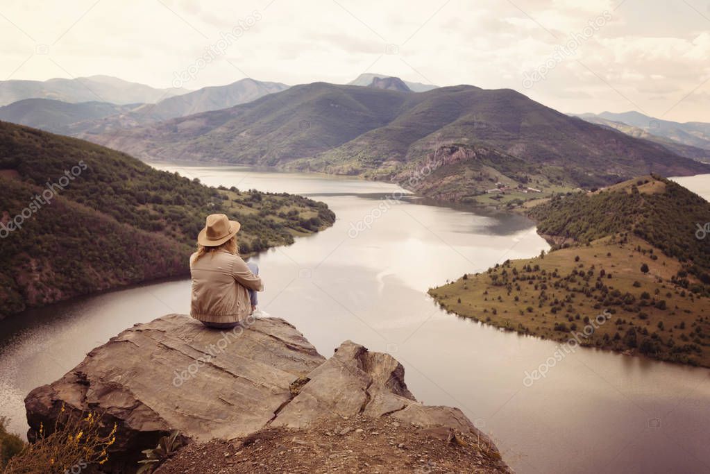 A hiking girl sits on the edge of the cliff and looking at the river and mountains. Location: the meanders of Arda river, Bulgaria