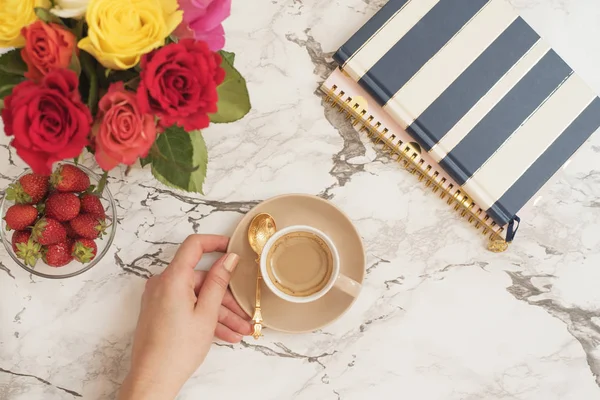 Feminine workplace concept. Freelance workspace in flat lay style with flowers. Woman hand holding cup of coffee. Blogger working. Top view, bright, pink and gold