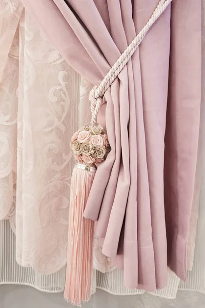 Part of beautifully draped curtain on the window in the room. Floral tassel tieback. Close up of piled curtain. Pink luxury curtain, home interior decor