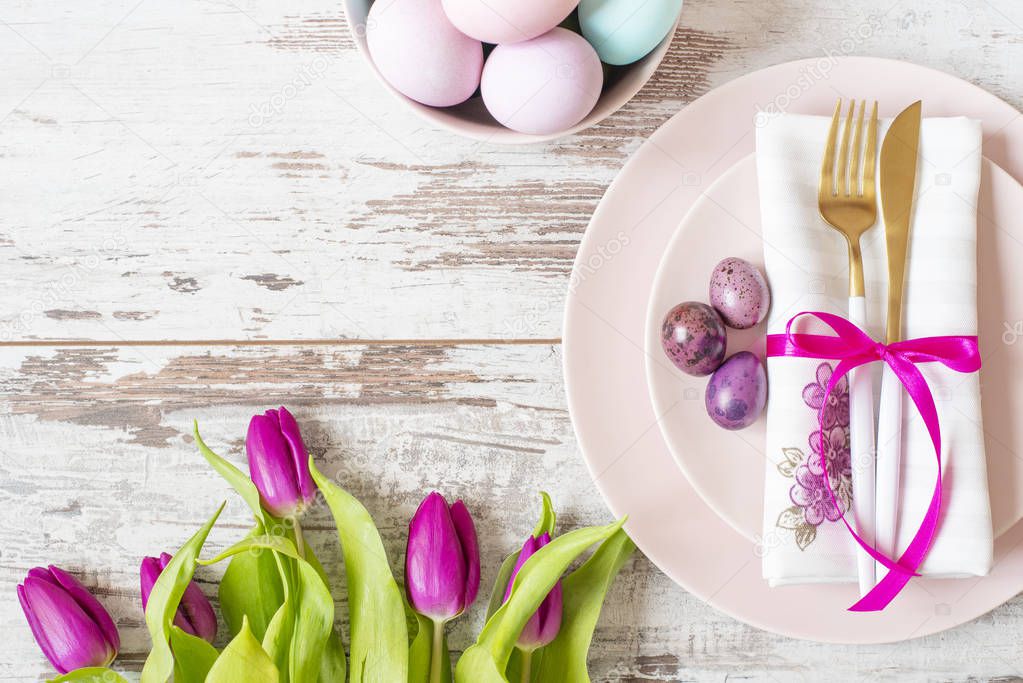 Easter table setting, flatlay with pastel colored Easter eggs, event decoration. Pink dishes and a gold fork and a spoon on a light rustic wooden background. Beautiful purple spring tulip flowers