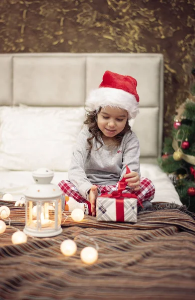 ¡Es Navidad! Feliz niña divertida sentada en la cama y abriendo el presente en la mañana de Navidad. Árbol de Navidad y luces alrededor Imagen De Stock