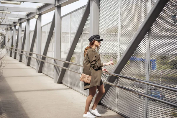 Modelo femenina de moda vestida casual en un lugar gruñón. Blogger de moda con camisa militar y sombrero en un puente peatonal Fotos De Stock Sin Royalties Gratis
