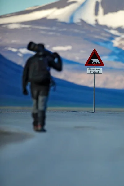 Photographer with big lens and road traffic sign with Polar bear. Man on the road with snowy mountain, travel in Svalbard in Norway. Danger big animal in the wild nature.