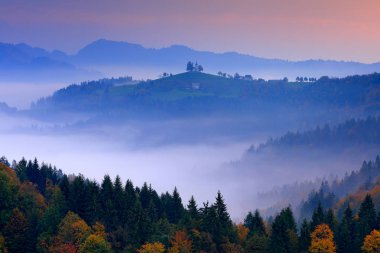 Alacakaranlık sis Slovenya ile. Aziz Thomas Kilisesi, Sveti Tomaz nad Praprotnim, Skofja Loka. Sisli Alpler ile orman, Slovenya, Europe seyahat. Mavi gökyüzü, sonbahar doğa ile güzel gündoğumu.