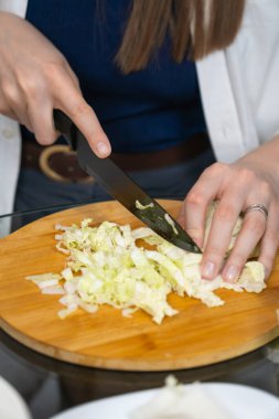 Female chef slicing fresh peking cabbage on a wooden cutting board, preparing ingredients for a healthy meal in a professional kitchen setting. Vertical photo