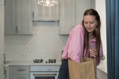Young woman with long brown hair, wearing a pink shirt and dark trousers, takes out groceries from a paper bag her modern kitchen seemingly pleased with her purchases