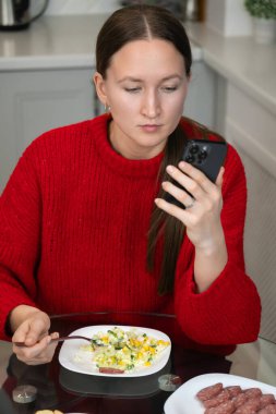 Sıcak kırmızı kazaklı genç bir kadın mutfak masasında akıllı telefonunu kullanırken sağlıklı bir öğle yemeğinin tadını çıkarıyor, sosyal medyayla meşgul oluyor ve mesajları karıştırıyor. Dikey fotoğraf
