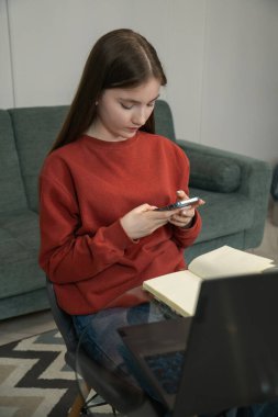 Focused young female student using a smartphone while studying or working remotely from home, surrounded by a laptop and books, creating a productive and comfortable workspace