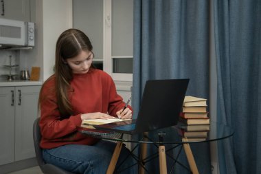 Focused teenage student student writing notes in notebook, using laptop and textbooks for online learning or homeschooling, demonstrating concentration and dedication to education