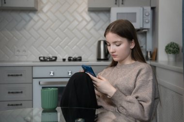 Young girl sits comfortably in her kitchen, savoring a mug of coffee while scrolling through her smartphone, enjoying a quiet moment of relaxation