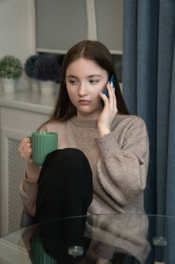 Teenage girl is sitting at a glass table, holding a green mug and talking on a blue smartphone, wearing casual clothes and having a serious expression. Vertical photo