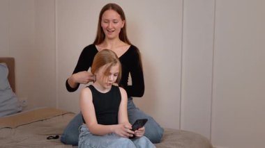 Sevecen anne, kızının saçlarını tararken genç kız yatakta dinleniyor, sabah aile içi iletişim ve bakım anında akıllı telefonları kaydırıyor.