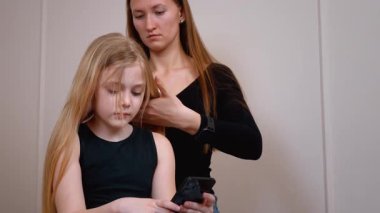 Sevecen anne, kızının saçlarını tararken genç kız yatakta dinleniyor, sabah aile içi iletişim ve bakım anında akıllı telefonları kaydırıyor.